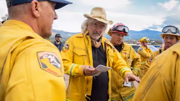 Firefighters in yellow gear gather outdoors as a man in a sun hat reads from a clipboard