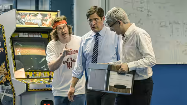 Doug (Matt Johnson) gestures beside two suited men examining a computer