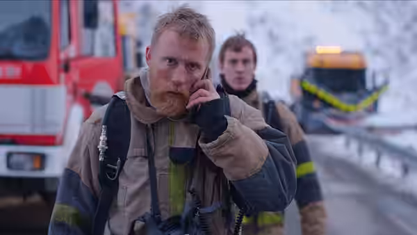 A bearded firefighter speaks into a phone beside emergency vehicles on a snowy road
