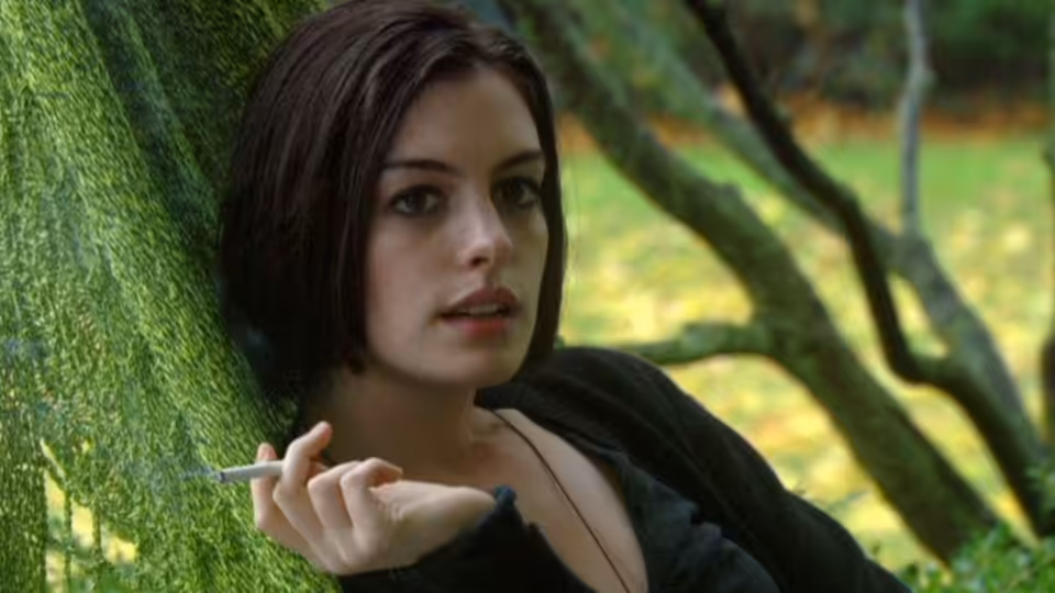 Kym (Anne Hathaway) leaning against a mossy tree while holding a cigarette