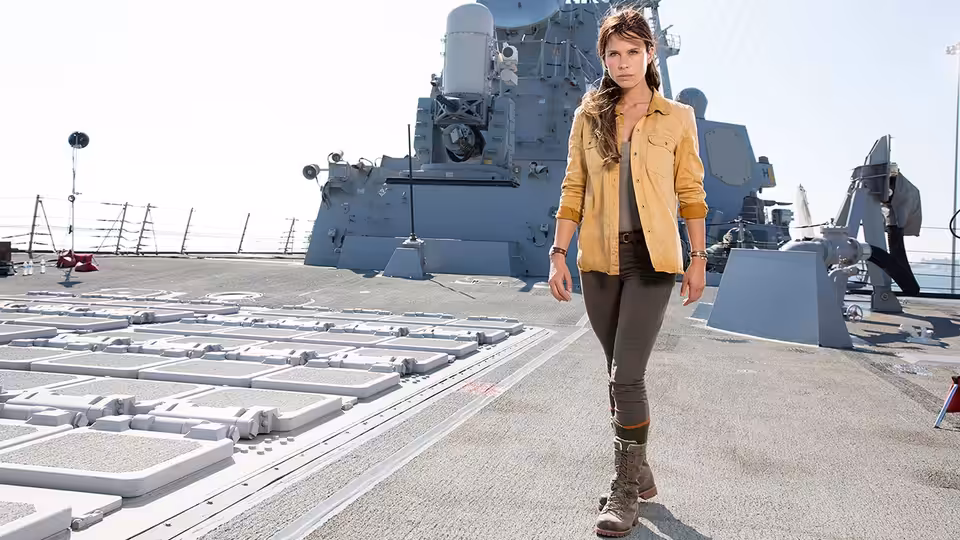 A woman in a tan jacket walks across the deck of a naval ship