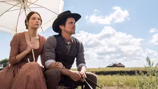 A woman and a man sit beneath an umbrella in a grassy field