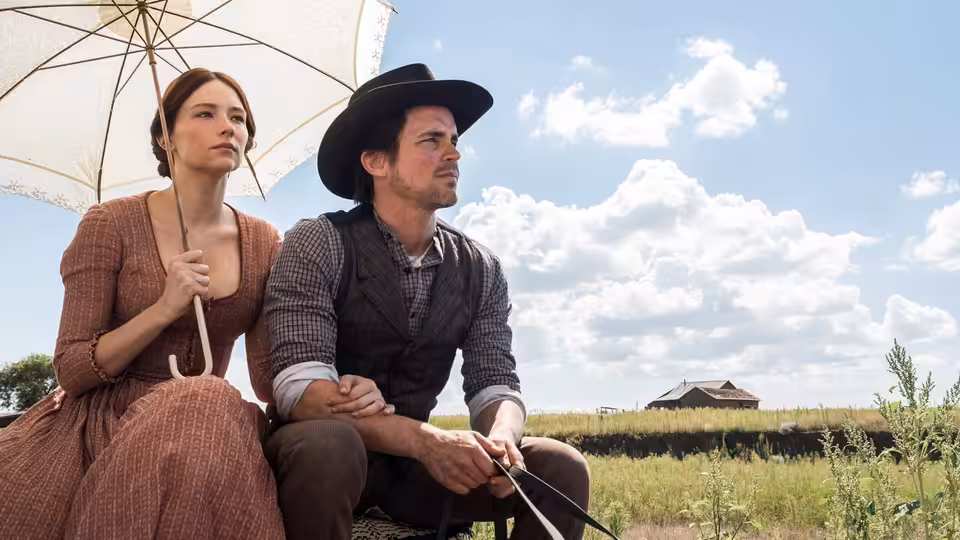 A woman and a man sit beneath an umbrella in a grassy field