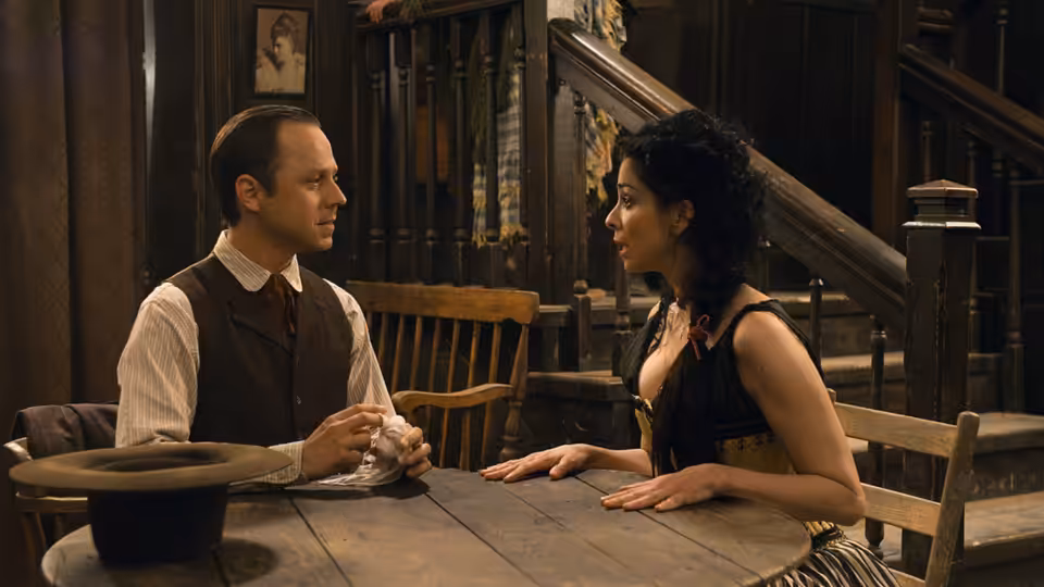 Albert and Anna sit facing each other across a wooden table indoors