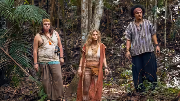 Three people stand among dense jungle foliage, dressed in casual outdoor clothing