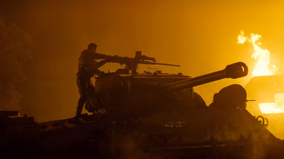 A soldier stands atop a tank silhouetted against a blazing sunset