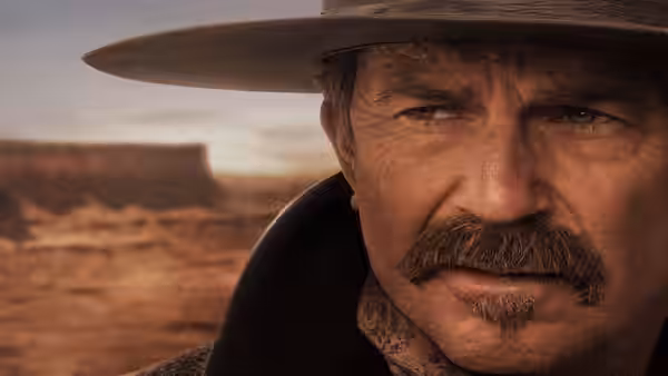 Hayes Ellison (Kevin Costner) stares ahead beneath his hat, with a frontier settlement behind him