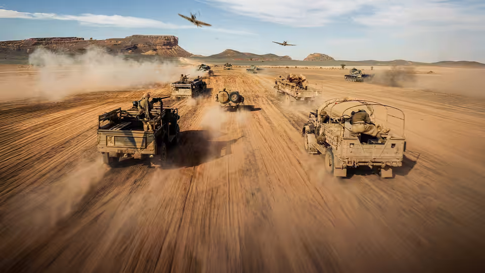 A convoy of military trucks crosses a desert airstrip beneath low-flying aircraft