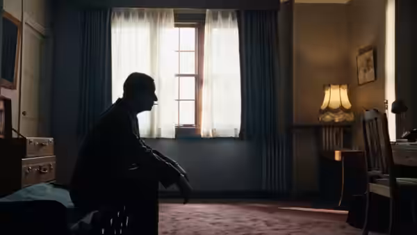 A lone man sits in silhouette beside a curtained window in a quiet room
