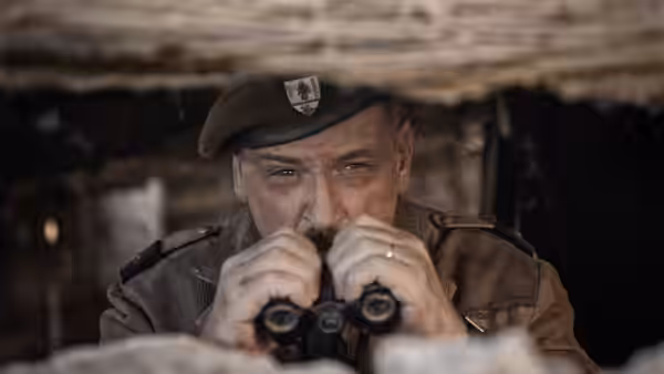 A beret-wearing officer peers through binoculars from a sheltered position