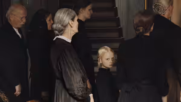 A blonde girl stands among adults gathered at the foot of a staircase