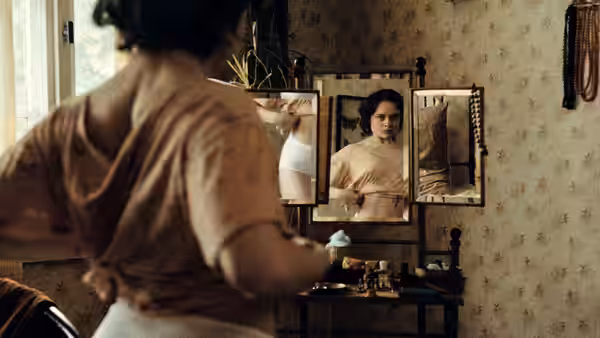 A woman adjusts her clothing before a mirror in a patterned room