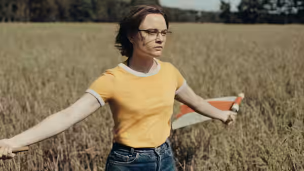 A bespectacled woman stands in a field with arms spread, holding a book