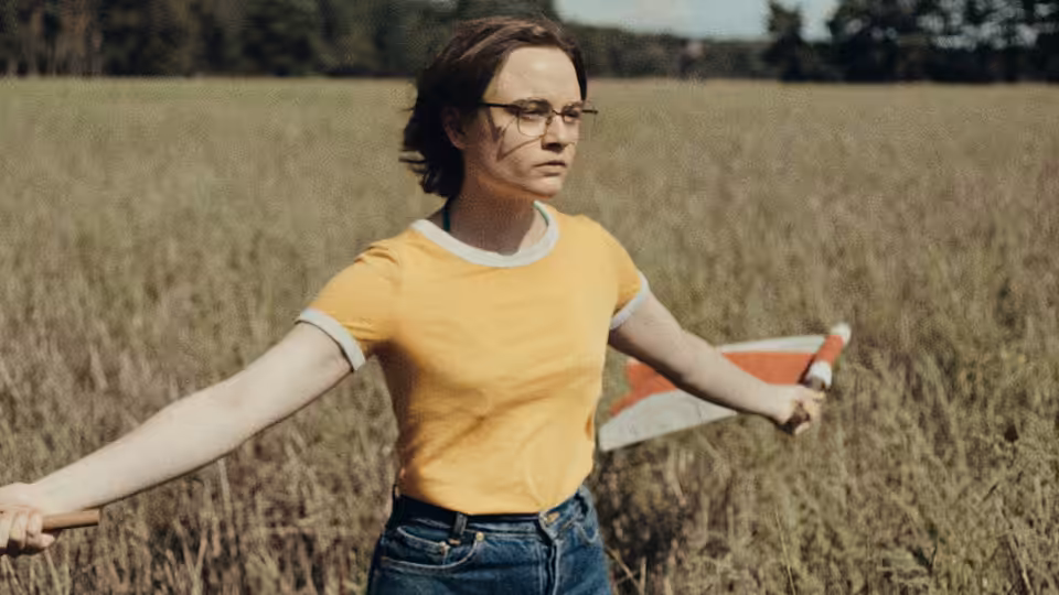 A bespectacled woman stands in a field with arms spread, holding a book