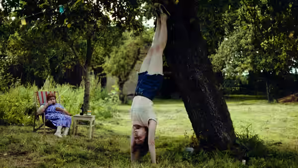 A girl does a handstand against a tree as a woman watches from a chair