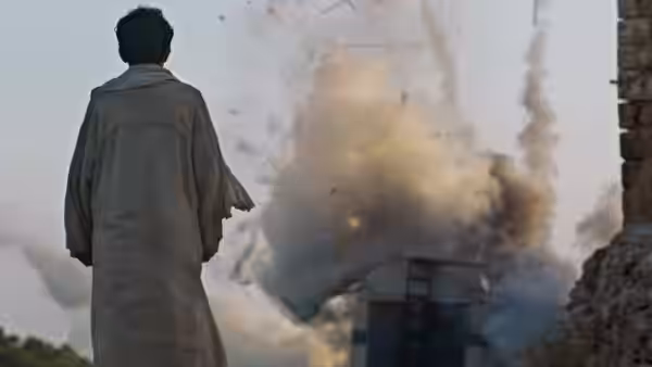 A robed man watches smoke billow above buildings from a street