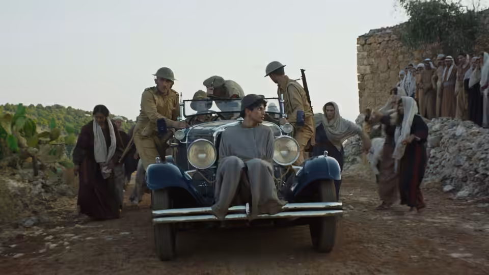 Villagers gather around a car carrying a seated man beside a stone building