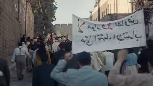 A crowd marches through a city street carrying a banner with Arabic writing