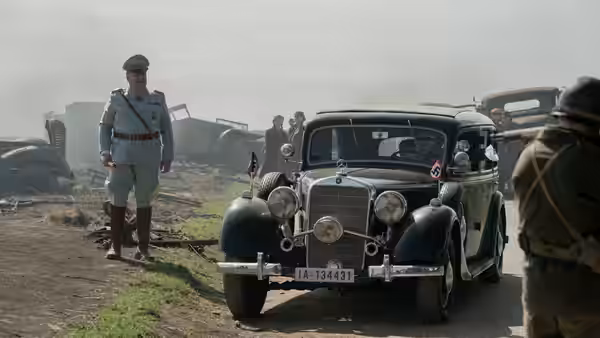 A uniformed officer stands beside a black car on a muddy military compound
