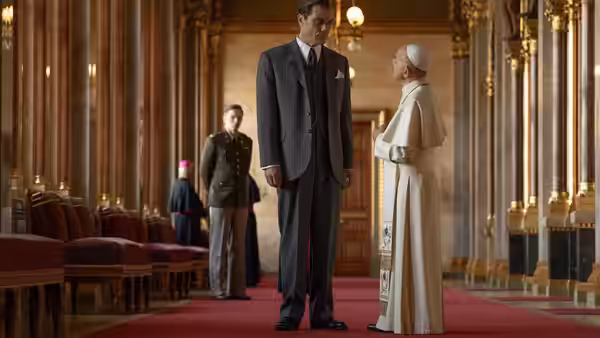 A suited official stands before a cleric in white robes inside an ornate hall
