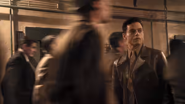 Douglas Kelley (Rami Malek) stands in a leather jacket as men hurry through a corridor