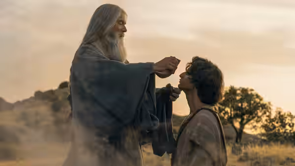 An elderly bearded man anoints a kneeling young man outdoors