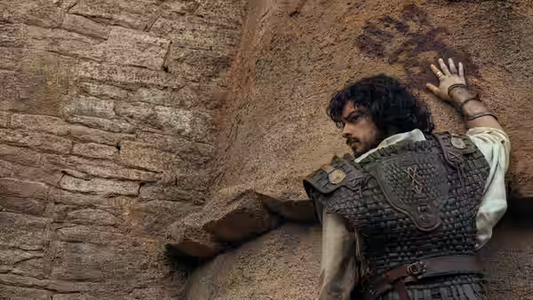 A young man in armor braces himself against a rough stone wall
