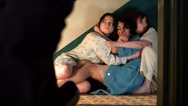 Three young women crouch together in a dim doorway, one holding another close