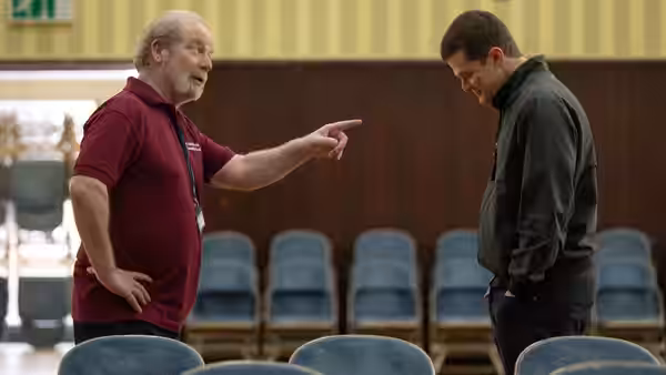 An older man points toward a younger man standing among chairs in a classroom