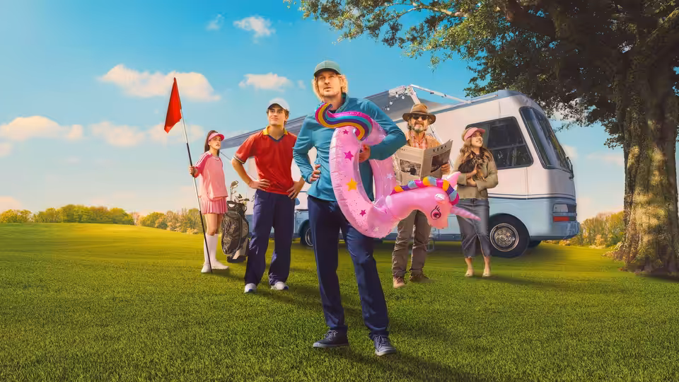 Pryce Cahill (Owen Wilson) holds a pink inflatable golf-course prop beside a camper van and group