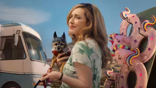 A woman holds a small dog beside a camper van and oversized pink inflatable props