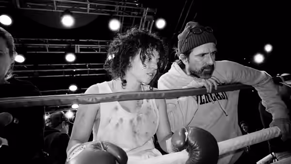 A boxer and beanie-wearing trainer lean over the ropes during a gym workout