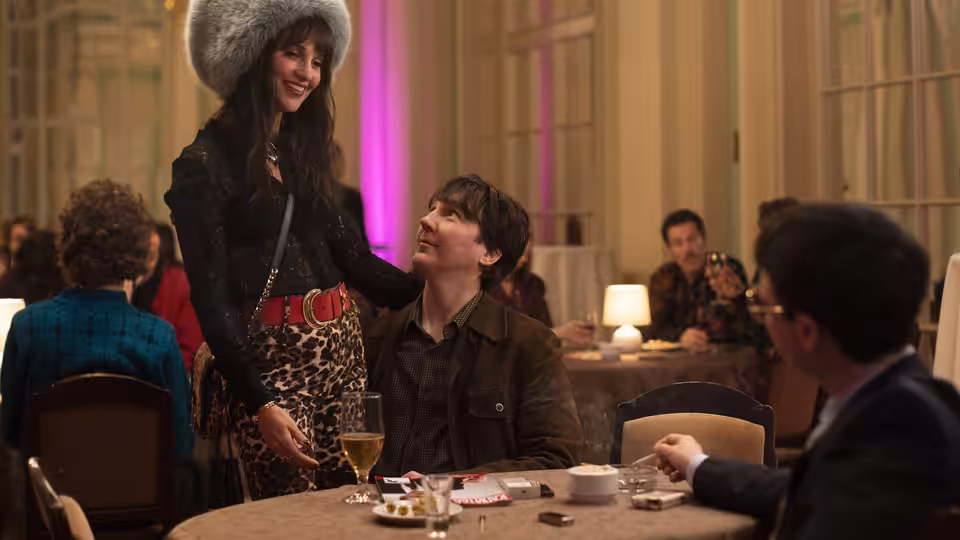 A woman in a fur hat stands beside a seated man at a restaurant table