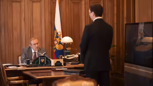 A bald official sits behind his desk as a man stands before him