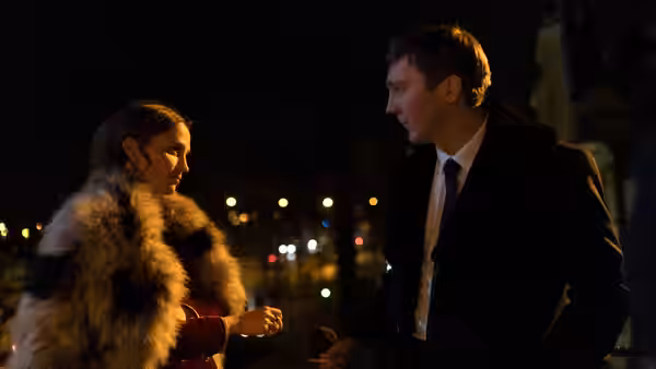 A woman in a fur coat talks with a suited man outdoors at night