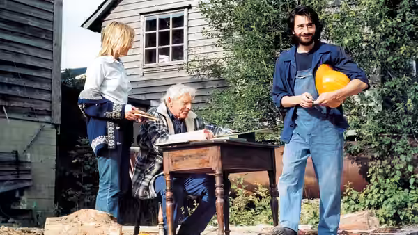 A bearded man holds a pumpkin beside a woman and an elderly man at a garden table