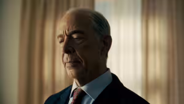A suited official stands in profile against a softly lit curtained window