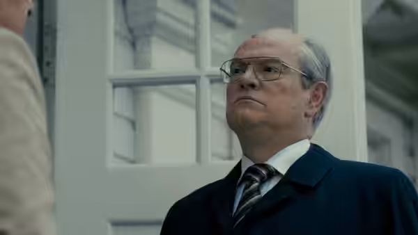 George Shultz (J.K. Simmons) looks upward in a suit and glasses