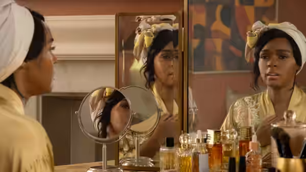 A woman in a headwrap studies her reflection at a vanity lined with bottles