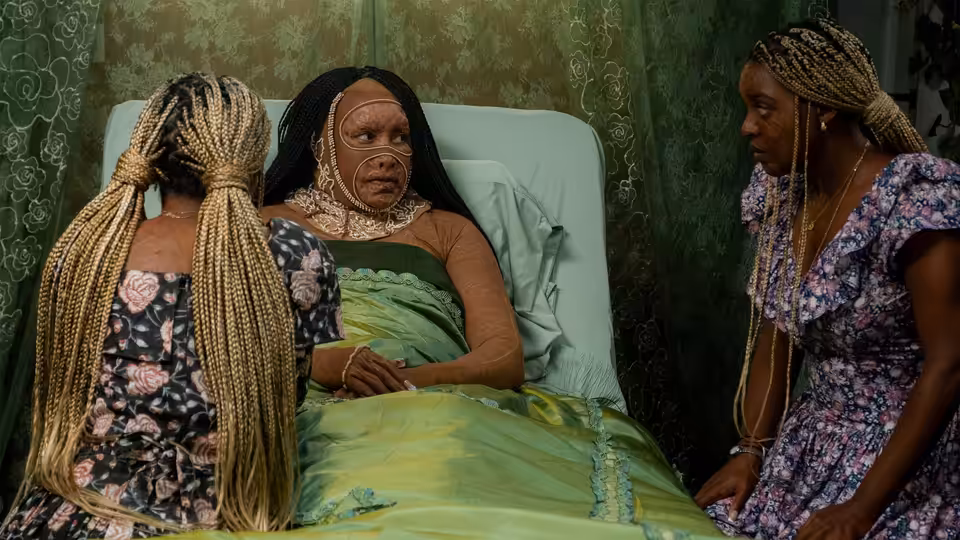 A woman lies in bed beneath a blanket as two women stand beside her
