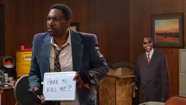 A man holds a handwritten sign reading “Here to Kill Me?” in an office