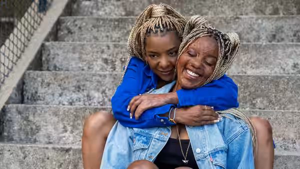 A seated woman smiles as a younger woman embraces her from behind on the stairs