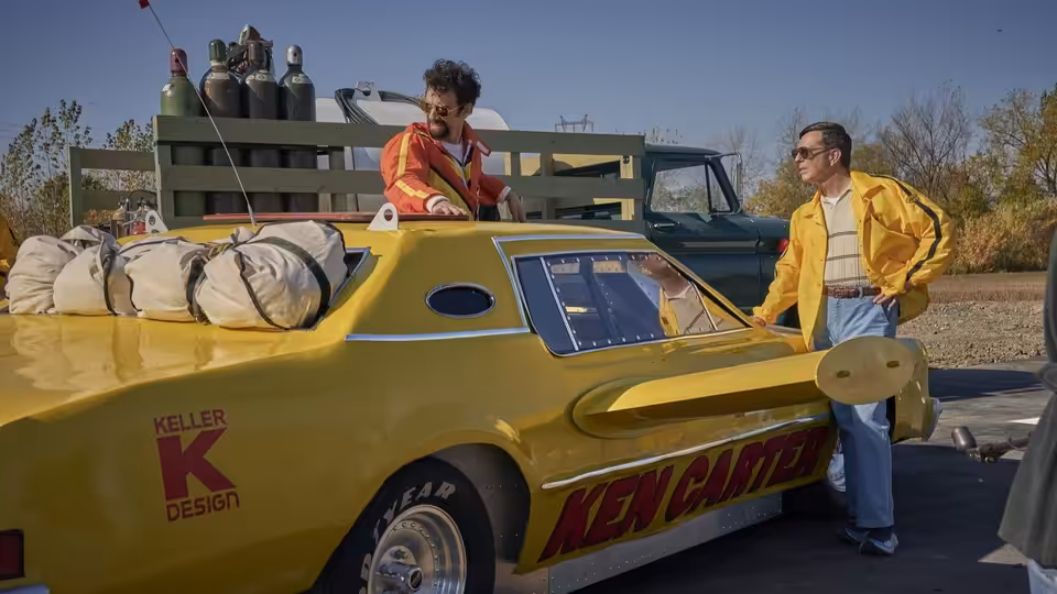 A man leans from a yellow stunt car as another stands beside it