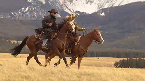 A bearded rancher and a blonde woman ride horses across a field