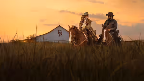 Two ranchers ride horses past a ranch building at sunset