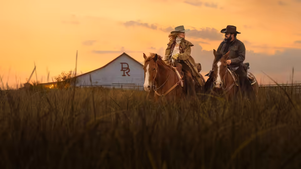 Two ranchers ride horses past a ranch building at sunset