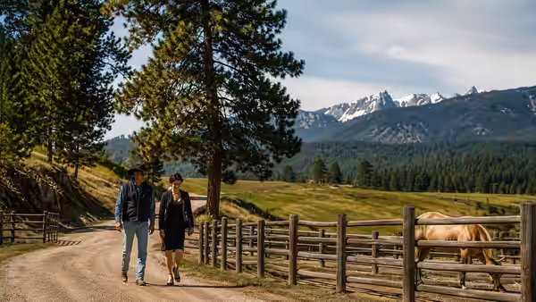 Two ranchers walk along a fenced paddock with mountains in the distance