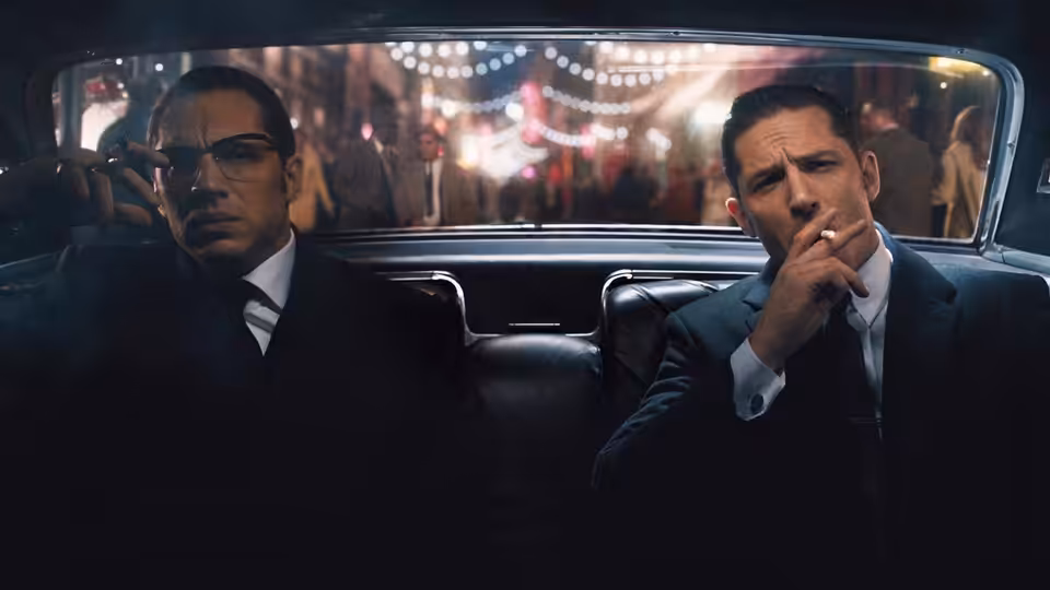 Ronald and Reginald Kray (Tom Hardy) ride in a car, one smoking in the back seat