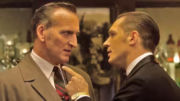 A suited man confronts an older man indoors, pointing a finger at his chest