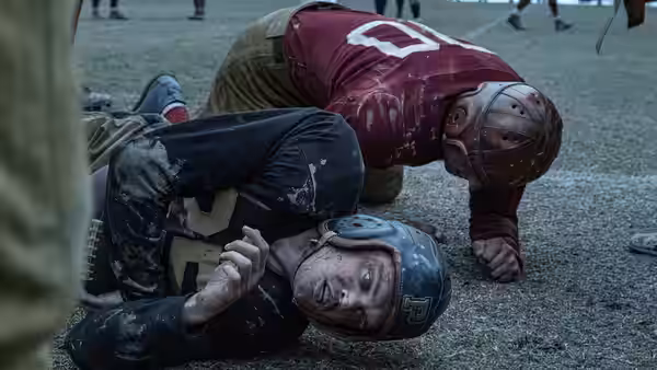A maroon-clad football player crouches over an opponent lying on the muddy field